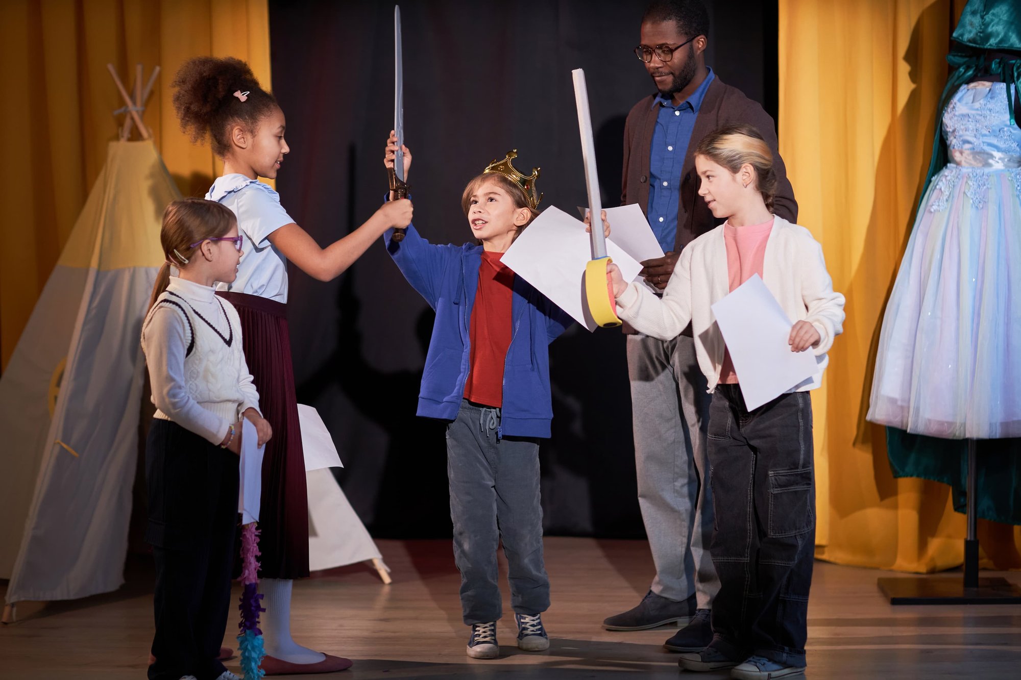Full length portrait of children rehearsing school play on stage in theater with little boy prince reciting lines
